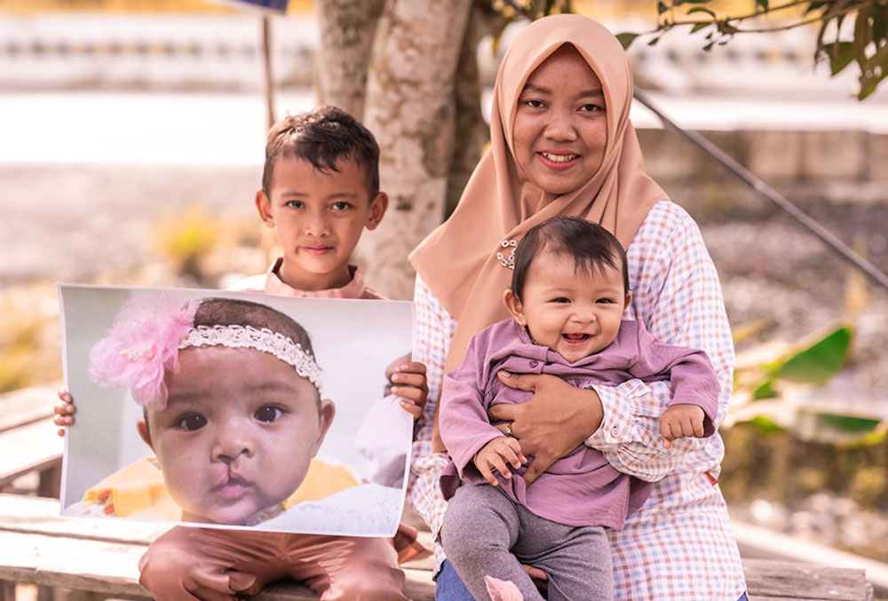 Divya mit ihrer Mutter Indri und ihrem Bruder, die lächelnd ein Bild von ihr vor der Spalt-OP halten